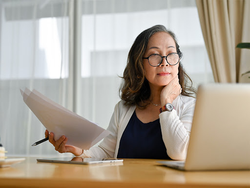 woman holding a piece of paper looking at her laptop screen deep in thought