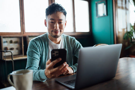 Confident young Asian man looking at smartphone while working on laptop computer in home office. Remote working, freelancer, small business concept.