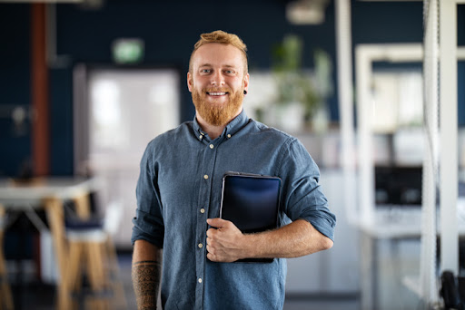 Portrait of a businessman with beard standing in office holding digital tablet. Confident male business executive in office looking at camera.